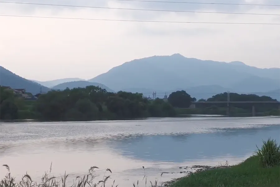 Calm river with grassy banks in the foreground and blue mountains in the distance under a pale sky.