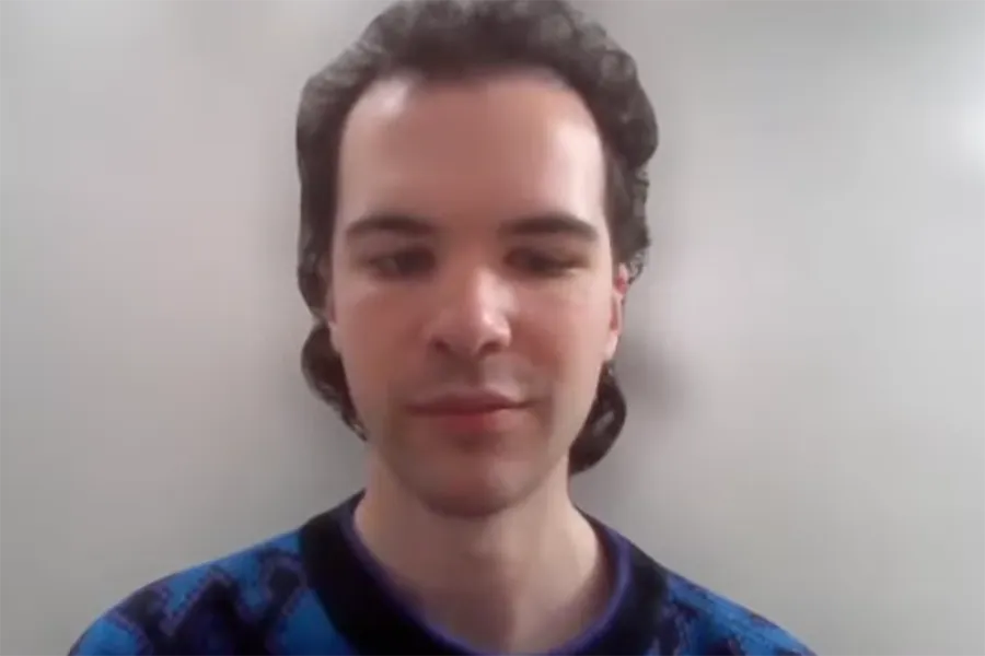 Young man with dark hair wearing a blue patterned sweater, seated against a light gray wall, looking slightly downward.