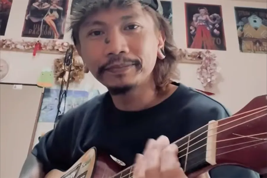 Man with a mullet and mustache playing an acoustic guitar indoors, facing the camera, with posters on the wall in the background.