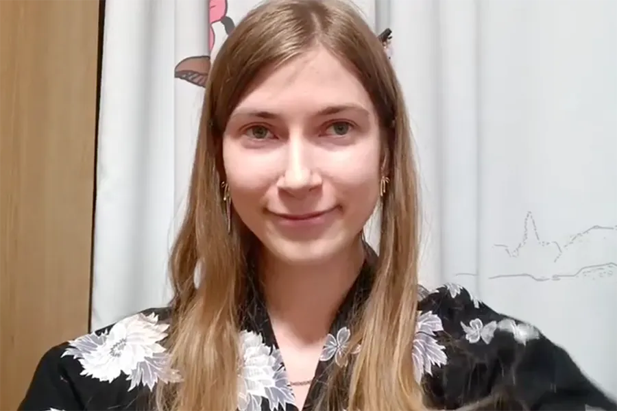 Smiling young woman with long blond hair wearing a black floral blouse, standing indoors by white curtains.