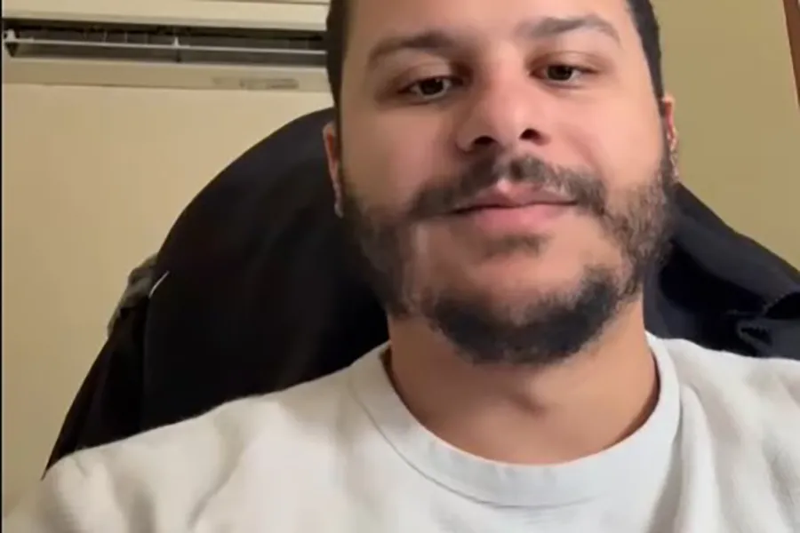Close-up of a man with short dark hair and a beard, wearing a light-colored shirt, looking at the camera during a video call indoors.
