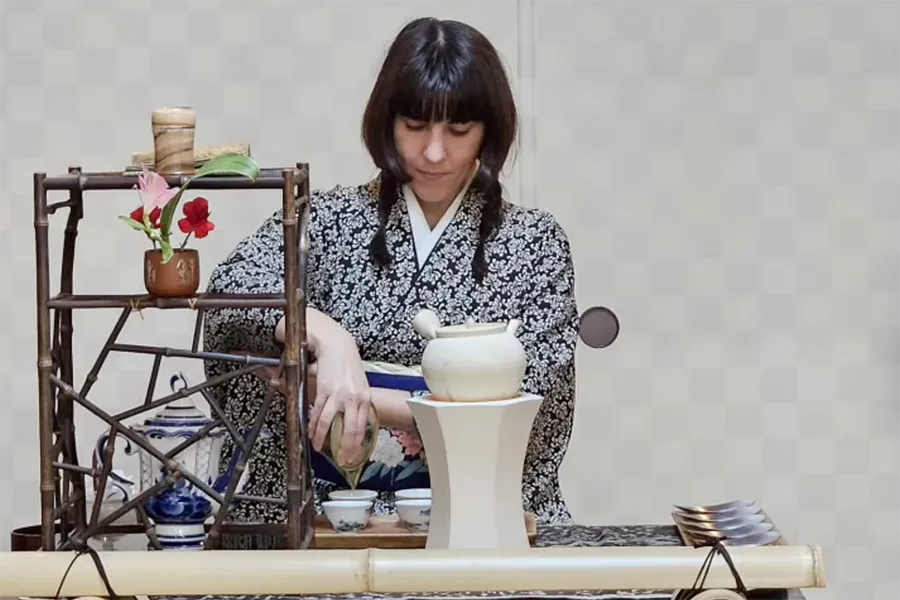 Woman in a patterned kimono performing a traditional Japanese tea ceremony with tea bowls and a bamboo rack nearby.