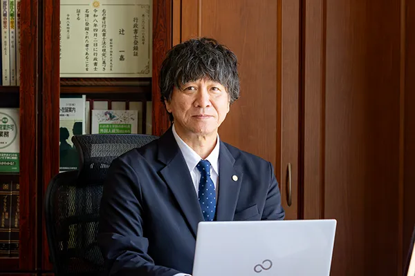 Professional man in a navy suit sits at his desk with a laptop in a wood-paneled office, facing the camera.